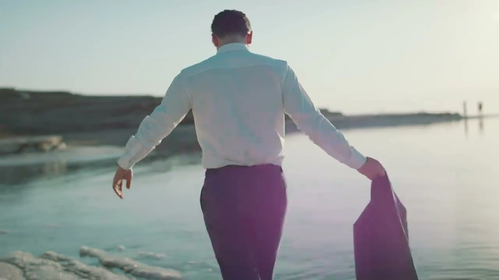 A man seen from behind beside sunlit water, carrying his jacket.