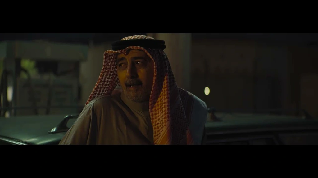 A man in a red-and-white keffiyeh speaking beside a car after dark.