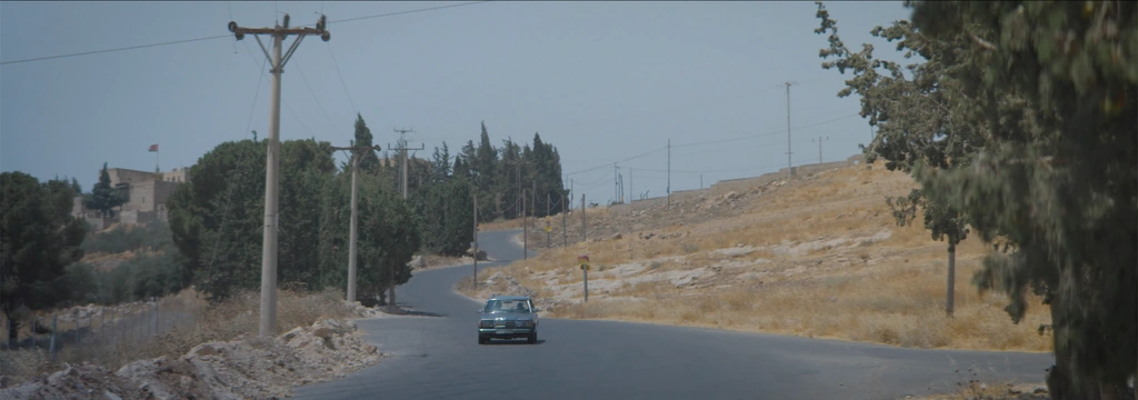 A car travelling along a winding road through a dry, tree-lined hillside.