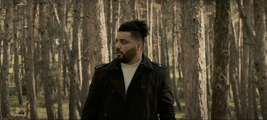 A man in a black jacket standing among tall pine trees.