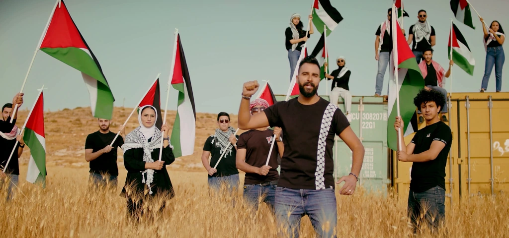 Toni Qattan performs in the foreground with flags and a container stage behind him.