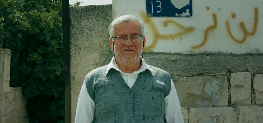 An older man stands beside a stone wall and a blue house-number sign.