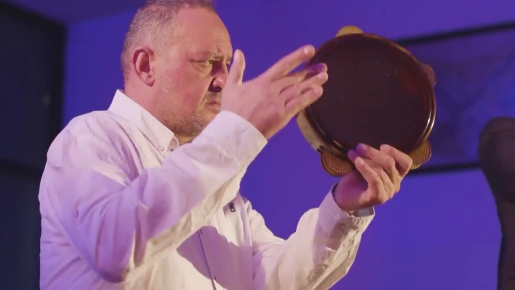 A percussionist raises his hand toward a frame drum in violet light.