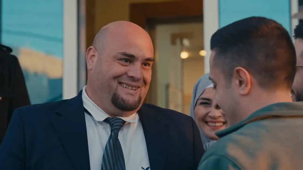 A smiling man in a dark suit greets another character outside an entrance.