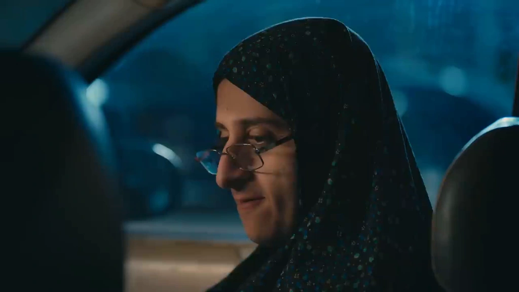 A character in a patterned headscarf sits inside a car under blue night lighting.