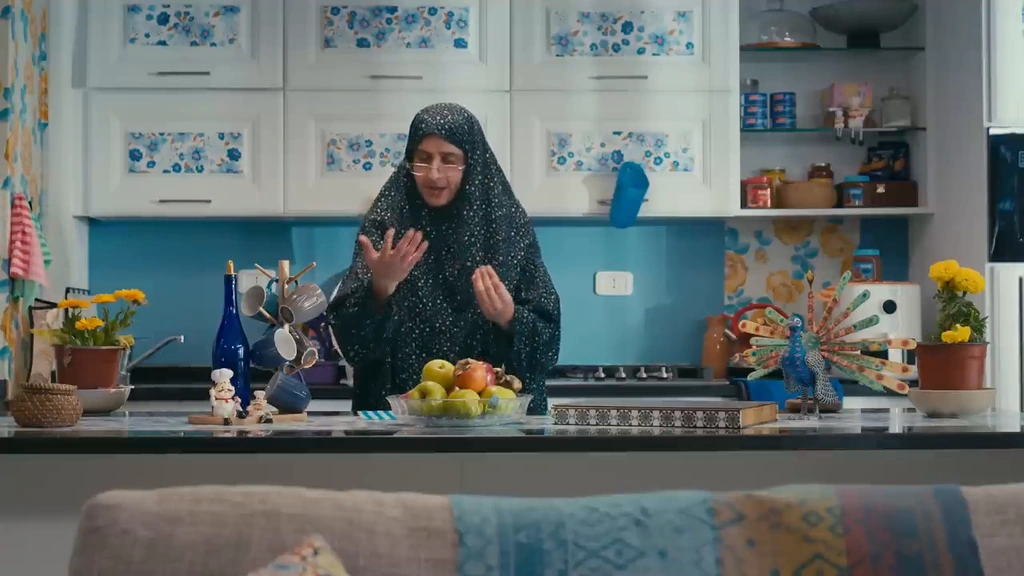 A character stands behind a kitchen island surrounded by fruit, patterned cabinets and blue walls.