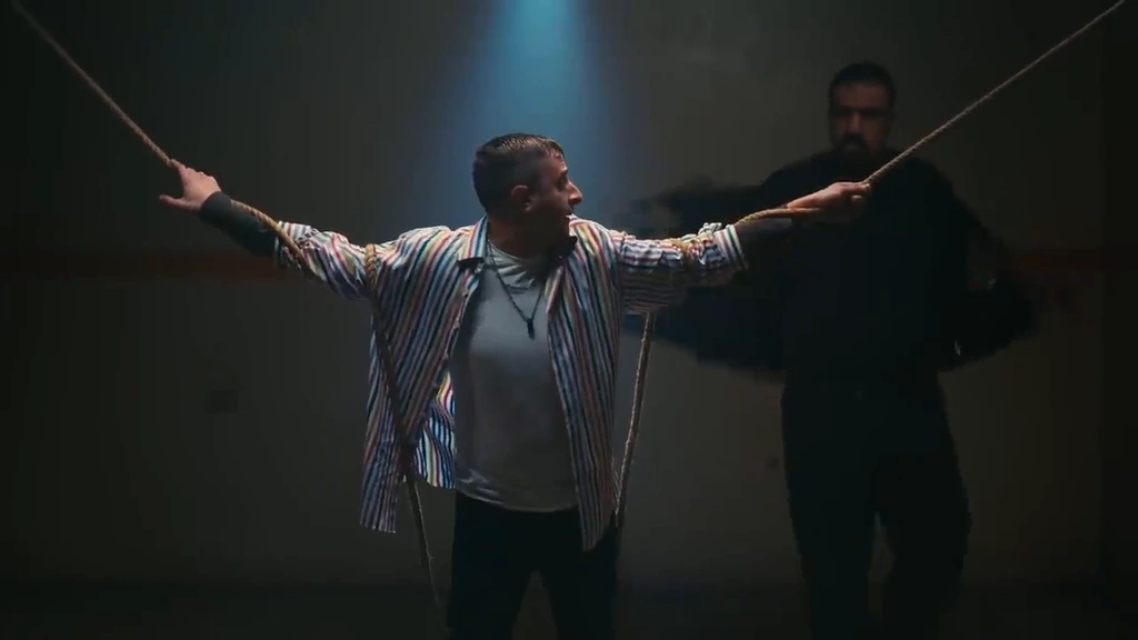 A man stands with his arms spread under a theatrical spotlight, with another figure in shadow behind him.