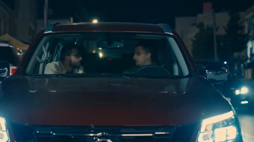 A front view of two men inside a car with its headlights glowing on a street at night.
