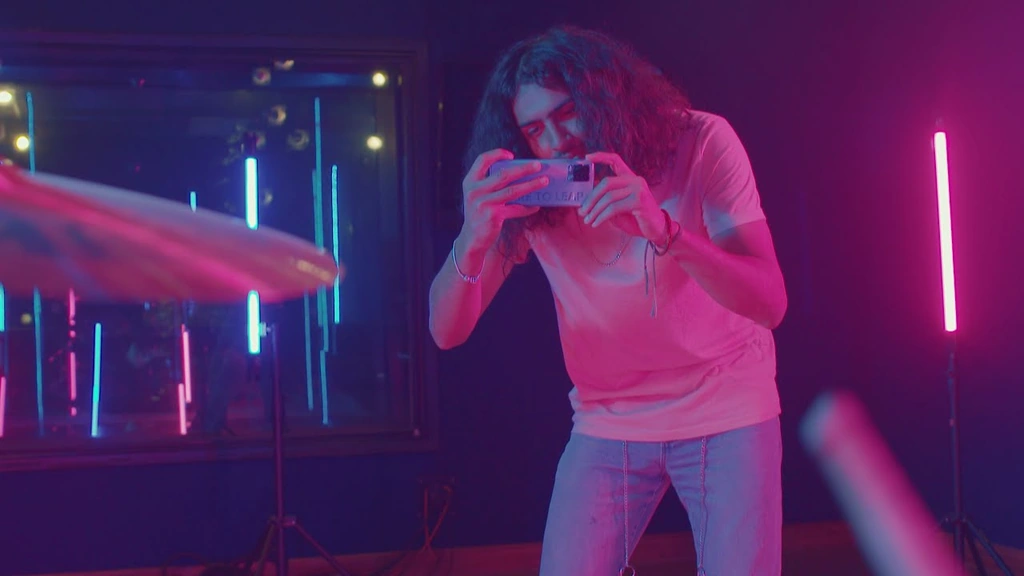 A person playing a mobile game beside a drum kit under magenta and blue light.