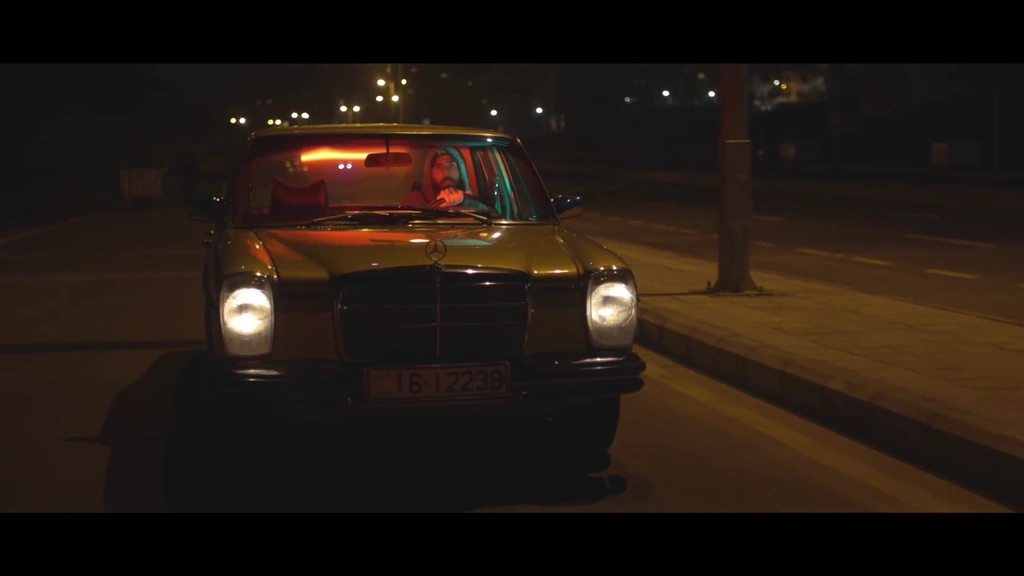 A classic car travels along a night road, its headlights facing the camera.