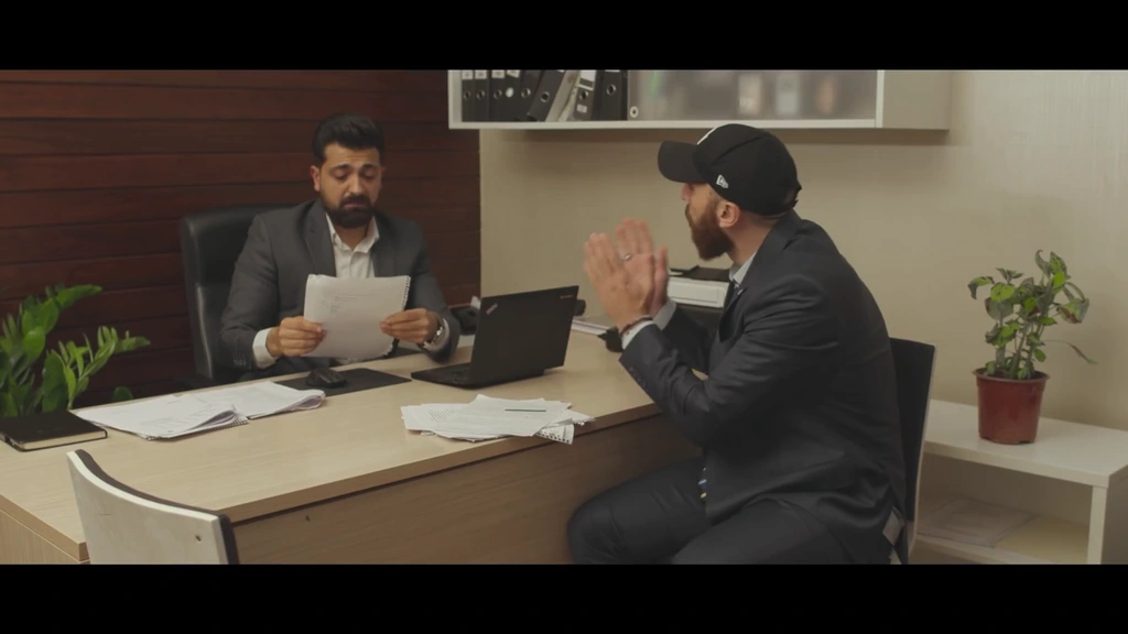 Two performers face one another across an office desk.