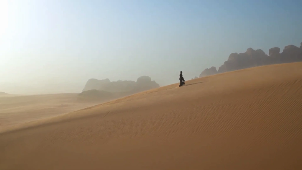A lone figure walks along the ridge of a sunlit dune beneath distant desert mountains.