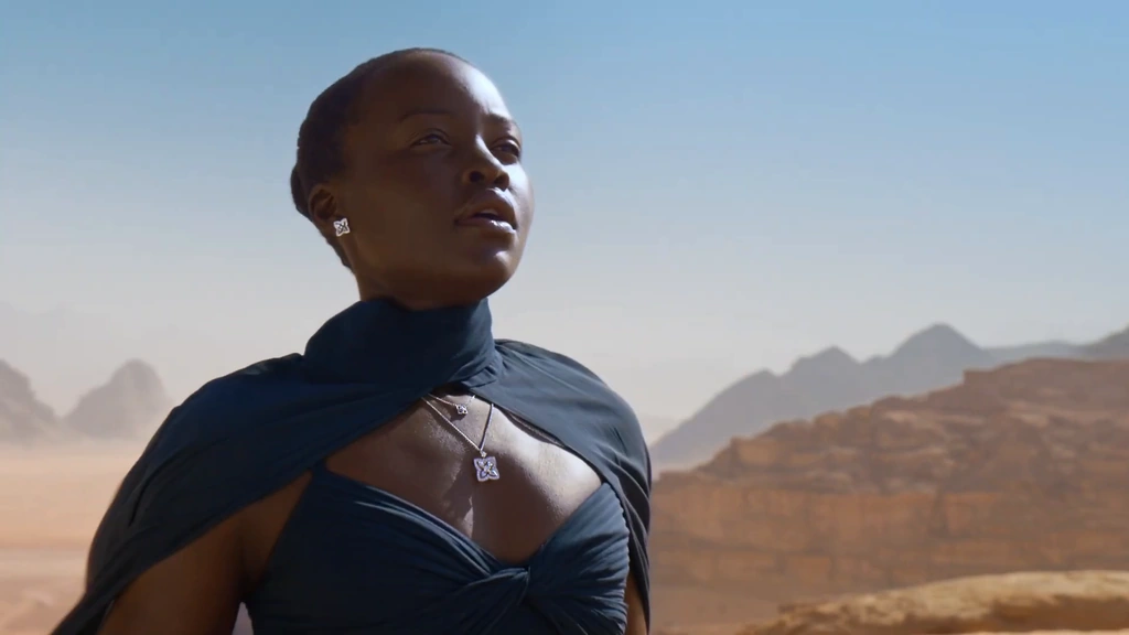 Lupita Nyong’o in a deep blue dress and diamond necklace against a sunlit desert.
