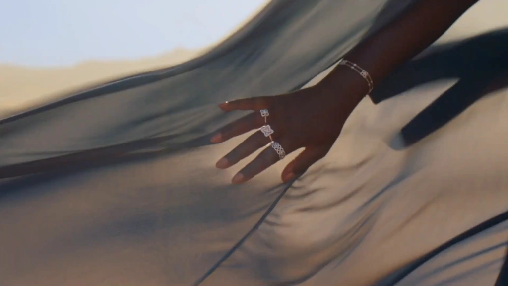 A hand wearing diamond jewellery lifts flowing blue fabric in desert sunlight.