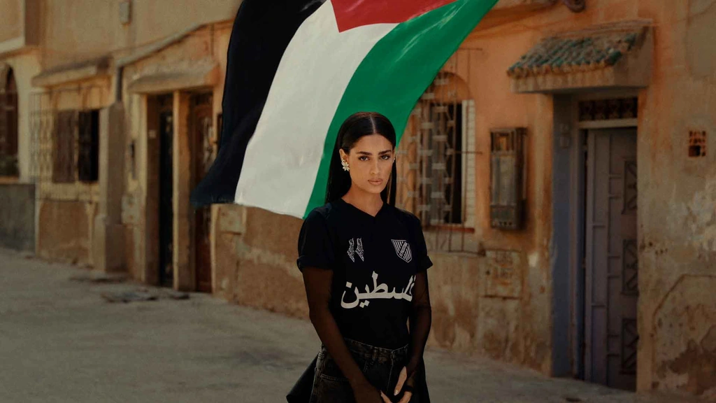 Zeyne stands on a stone street beneath a Palestinian flag.