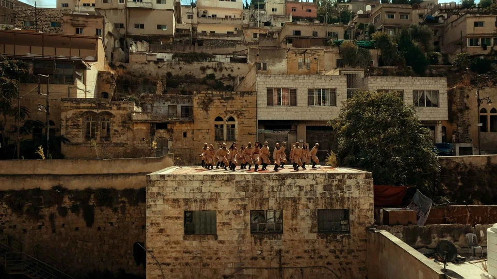 A line of dancers on a stone rooftop surrounded by hillside buildings.