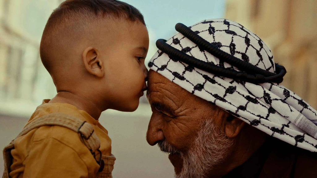 A young child kisses the forehead of an older man wearing a keffiyeh.