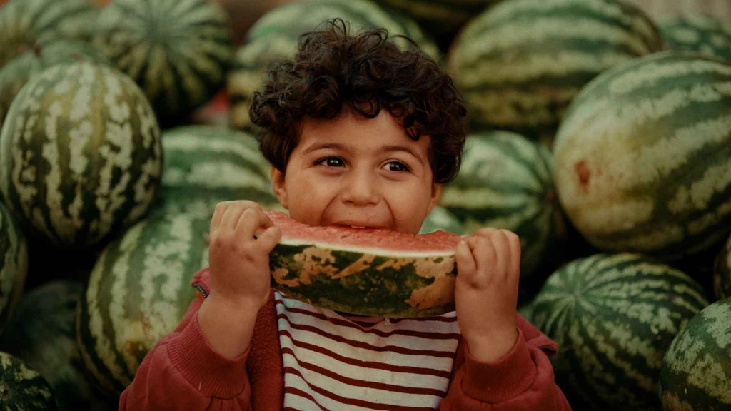 A child smiles while holding a slice of watermelon in front of a market display.