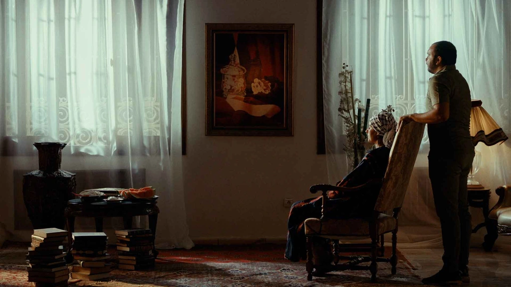 Two men in a quiet interior lit by large windows and sheer curtains.